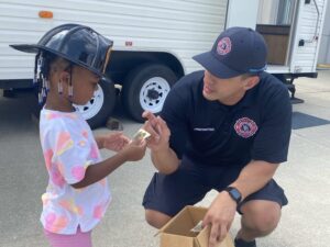 A firefighter giving a badge sticker to a child wearing a play firefighter hat.