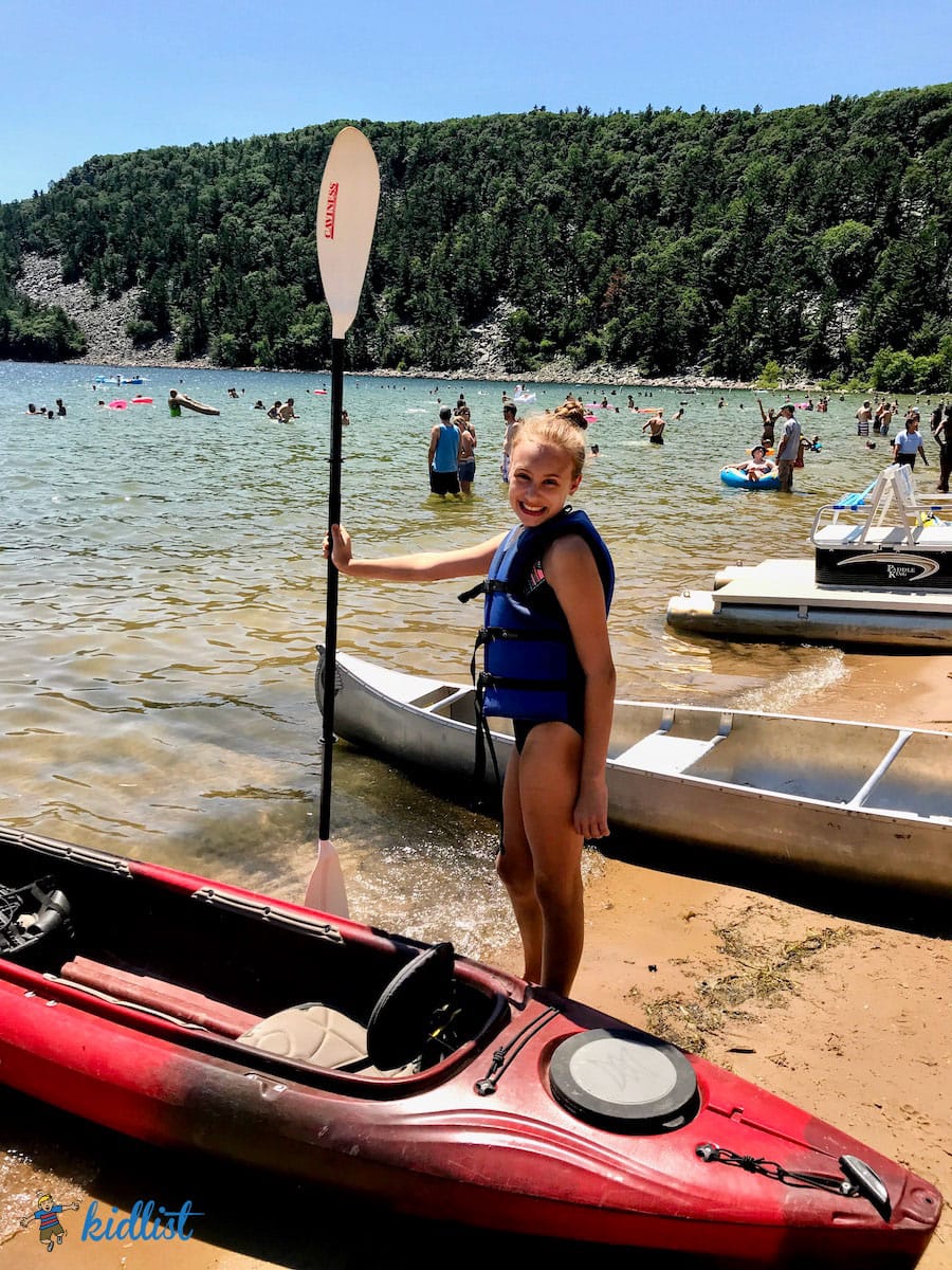 Girl in a lifejacket smiling and holding an oar next to a kayak on the beach.