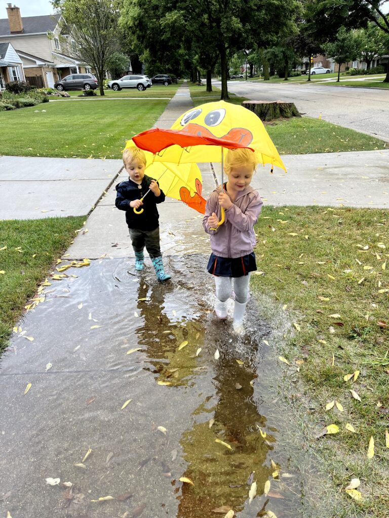 Two young children splashing in a puddle on a rainy day.