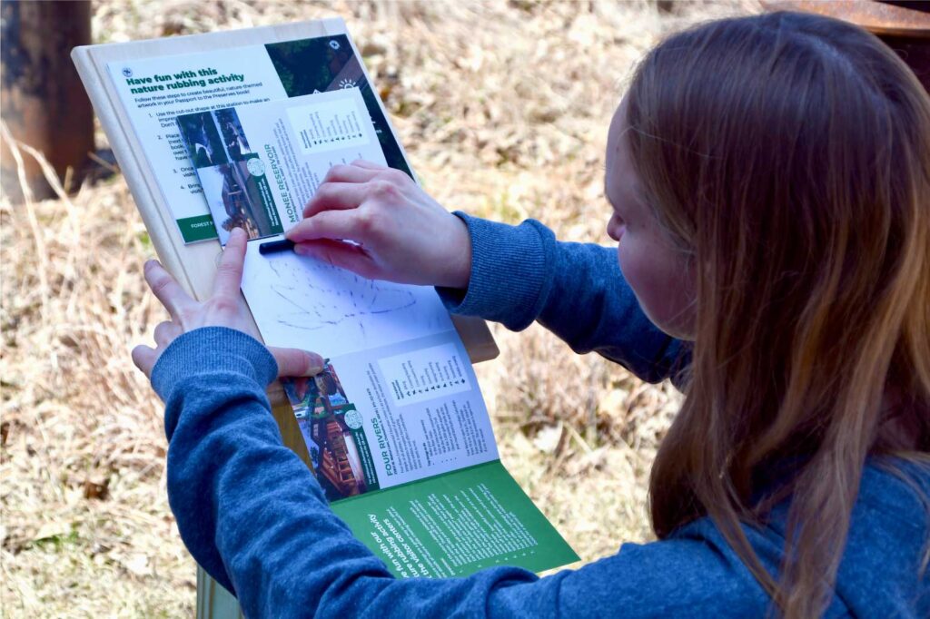 A person takes a rubbing of a leaf from a placard, using a forest preserve passport brochure.