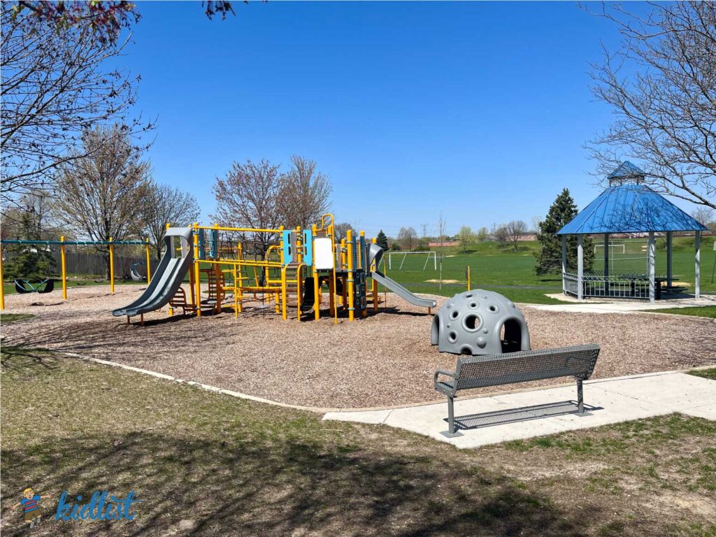 Park with a bench, woodchips, play structure, and gazebo near soccer fields.