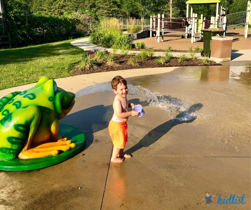 Child at a splash pad with a colorful frog fountain.