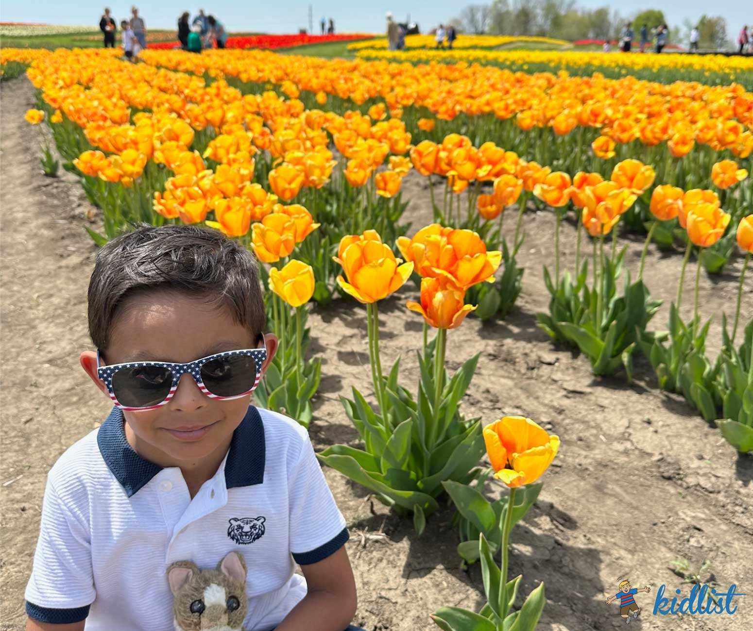 A child with sunglasses holding a stuffed animal stands smiling in a field of tulips.
