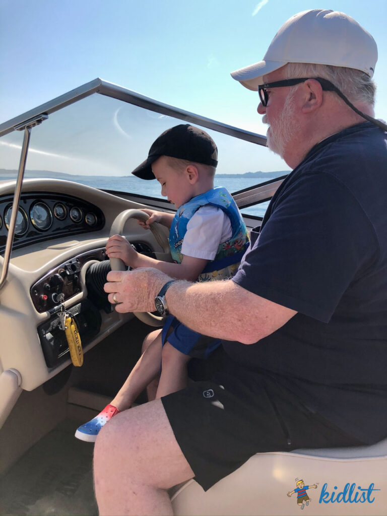 A boy and his grandpa on a boat. The boy is on his Grandpa's lap as he lets him steer the boat
