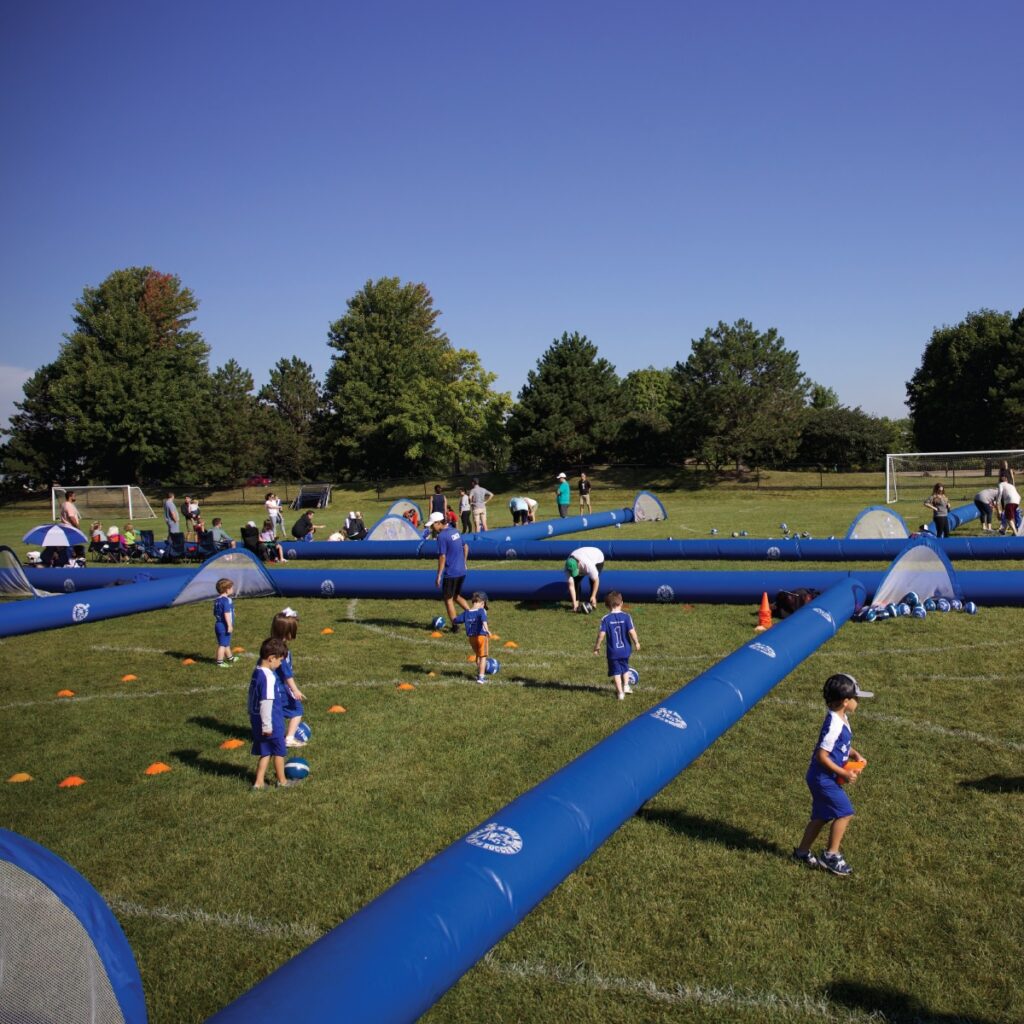 Kids in blue Lil Kickers uniforms practicing soccer on a grassy field with inflatable dividers and mesh goals.