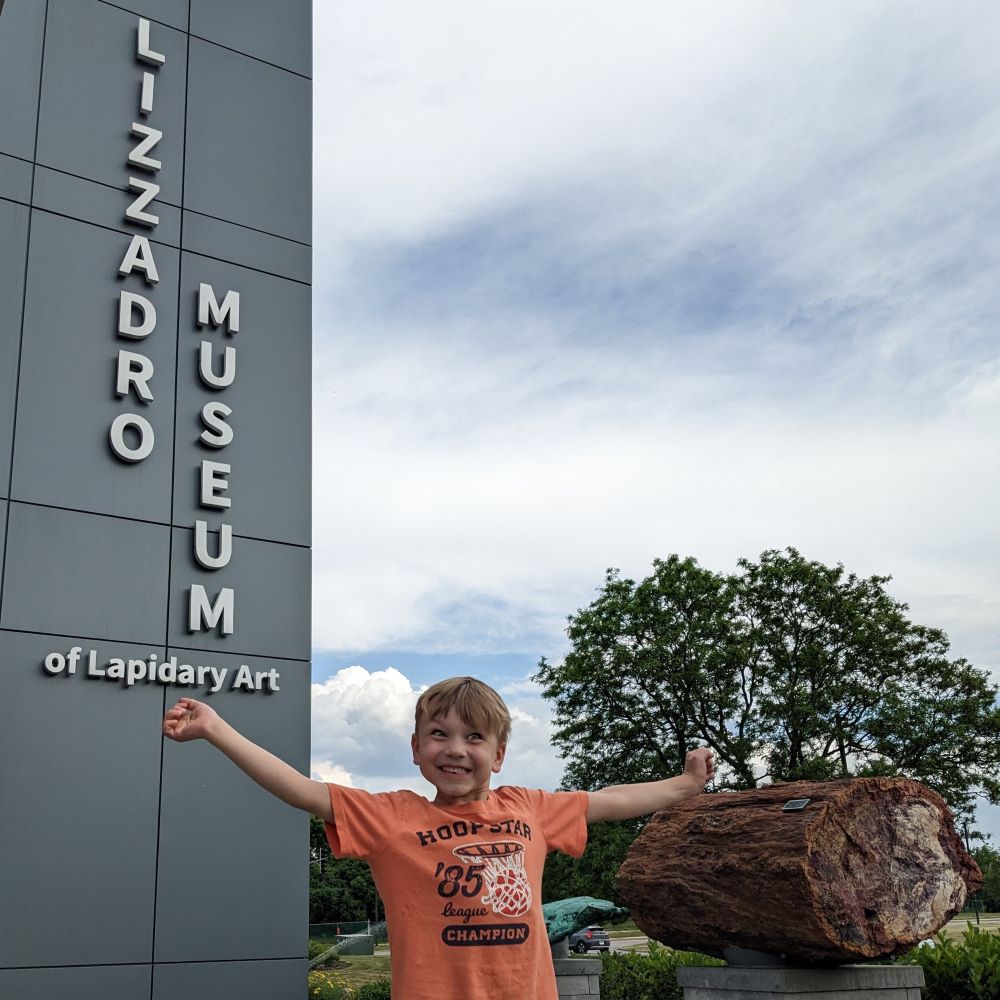 Child grinning and stretching their arms in front of the sign for the Lizzadro Museum of Lapidary Art