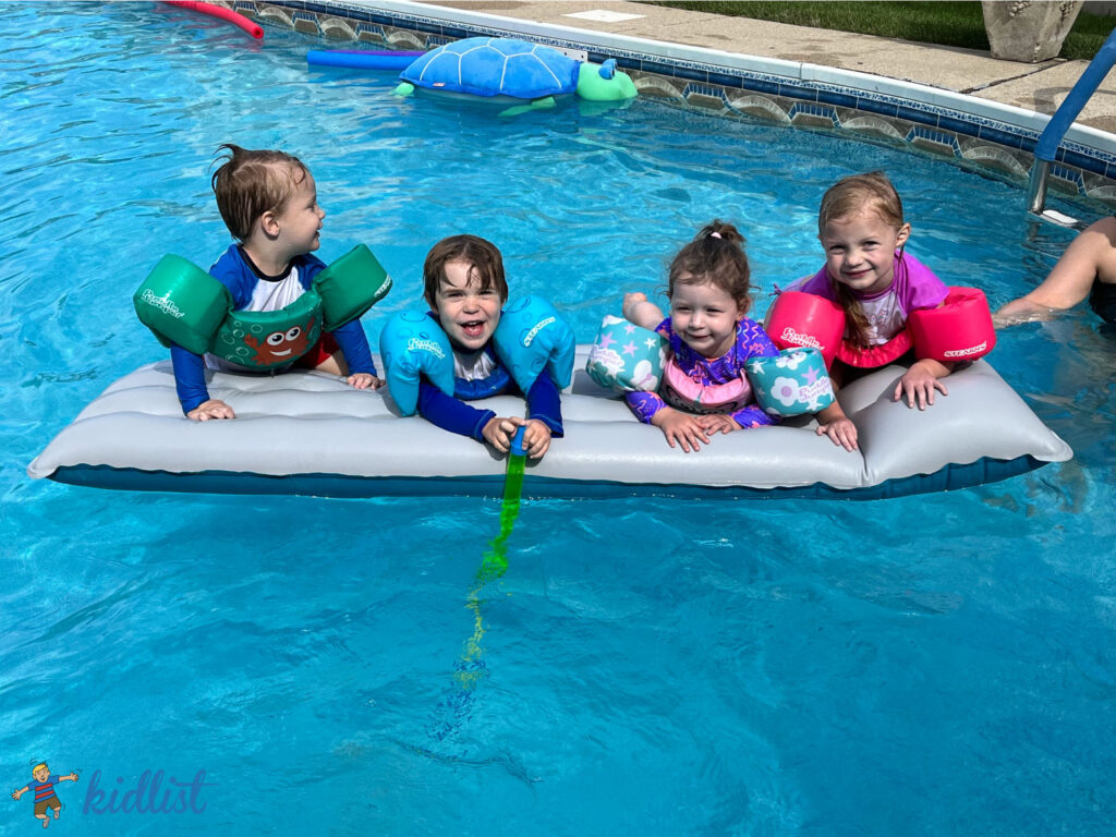 four kids laying on an inflatable float in a bright blue outdoor pool with crystal clear water