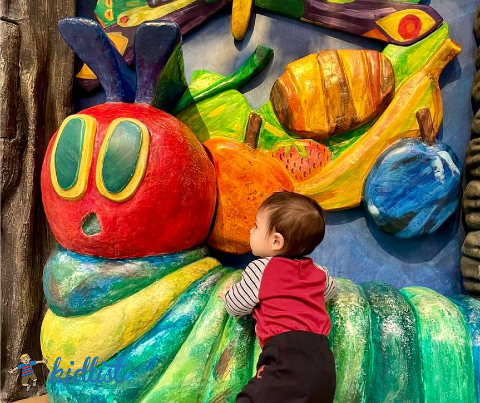 A toddler holds onto a large, colorful sculpture of The Very Hungry Caterpillar from the Eric Carle children's book. The photo is from the Schaumburg Township Library's children's area.
