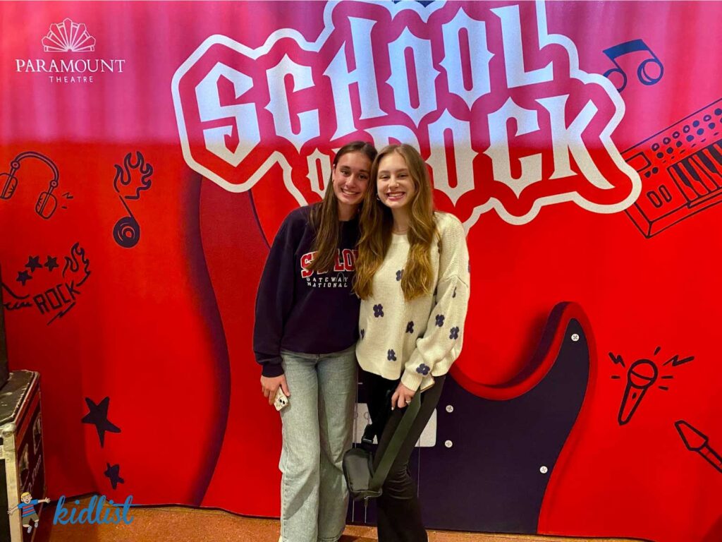 two sisters standing in front of a red background with the School of Rock logo on it