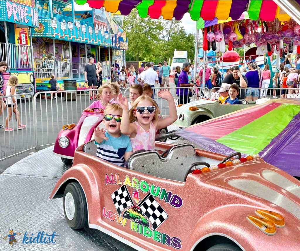 Kids smiling and waving on a car-themed merry-go-round ride at a summer festival.