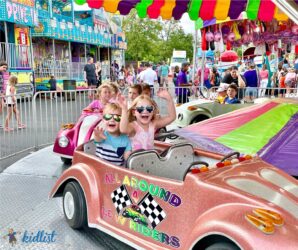 Kids smiling and waving on a car-themed merry-go-round ride at a summer festival.