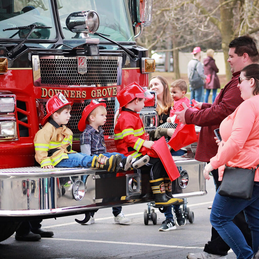 Kids dressed up as firefighters pose for a photo on the front bumper of a fire truck.