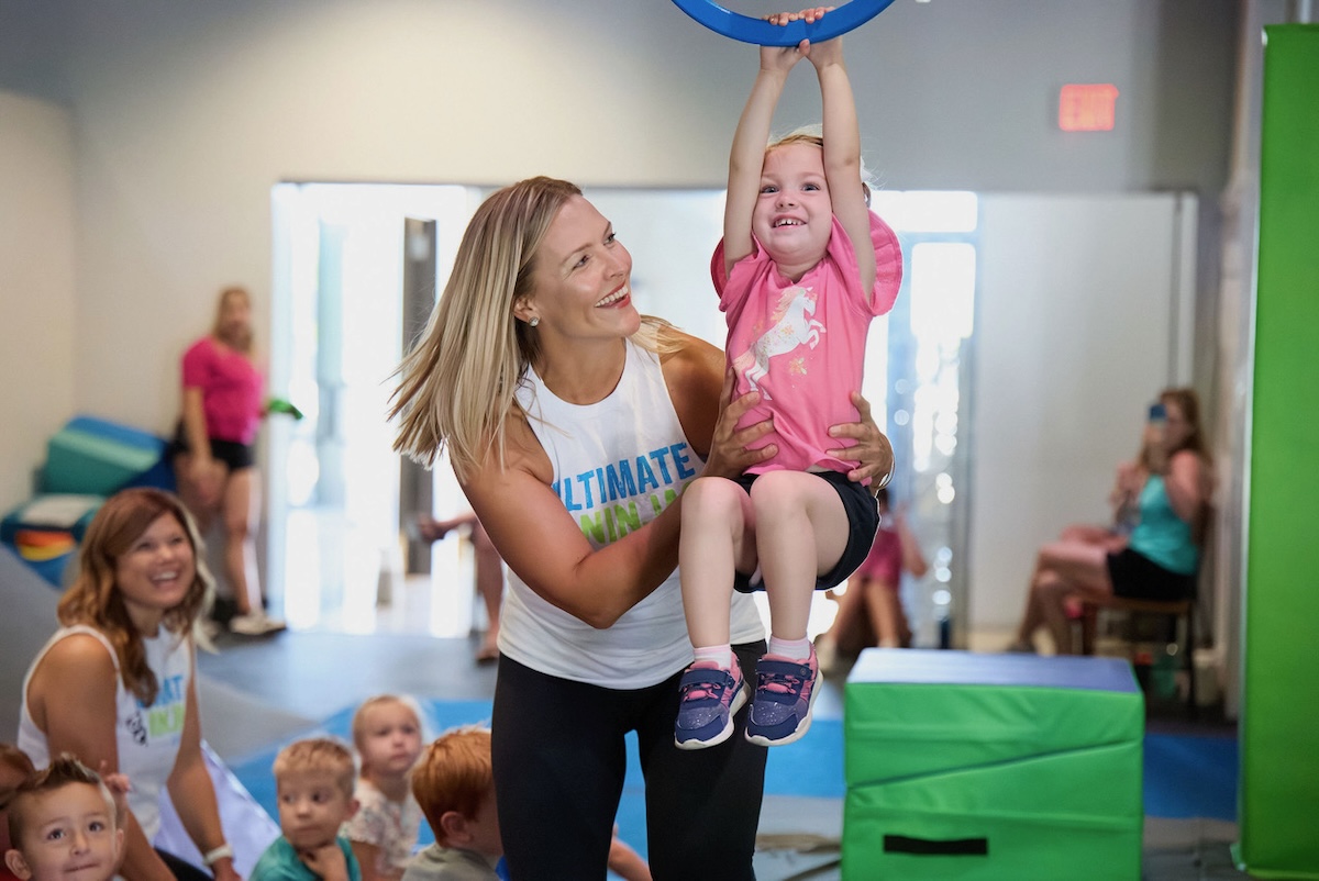 A smiling coach helps a young girl swing on a ring, with other children and soft tumbling mats in the background.