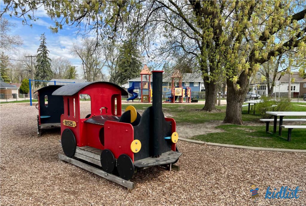 Toddler-friendly train play structure at a larger playground with a fence surrounding the big area. Woodchip play surface.