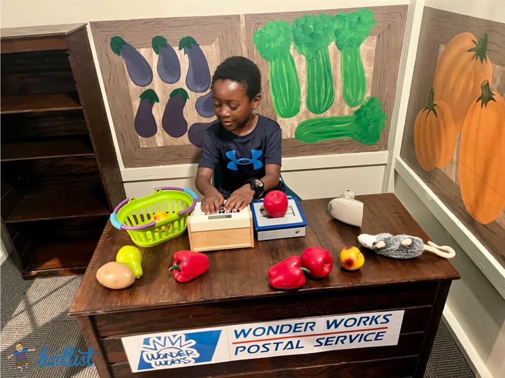 A child weighing objects on a play scale at a station labeled Wonder Works Postal Service.