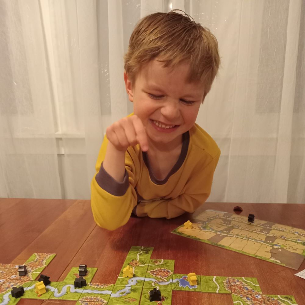 A kid pointing down at a game of Carcasonne and grinning.